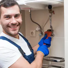 Uśmiechnięty hydraulik w niebieskich rękawiczkach naprawia bojler kluczem nastawnym.